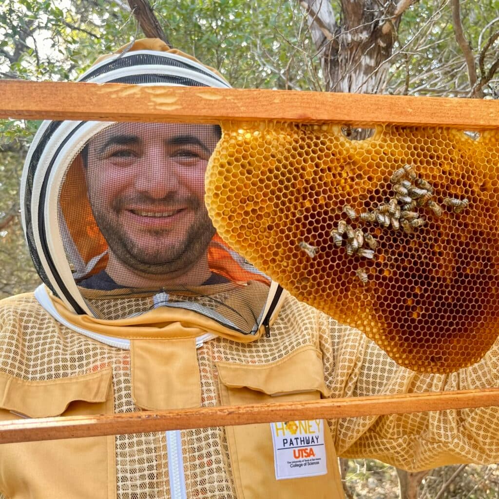 Dr. Ferhat Öztürk in a beekeeper suit holding a honeycomb frame